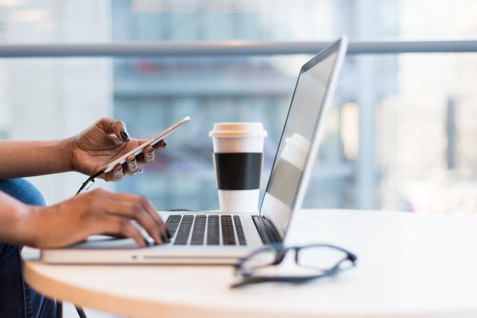 mãos segurando no celullar e teclando no notebook com copo de café ao fundo