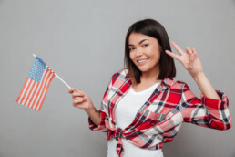 mulher sorrindo com a bandeira dos estados unidos na mao e fazendo dois nos dedos
